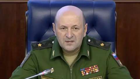 AP Igor Kirillov, bald and wearing blue military overalls, speaks into a microphone during a press conference.