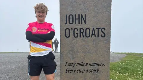 Rafe Colman-Chadwick A man stands next to the John O'Groats sign that says 'Every mile a memory, every step a story'. He has pale skin, blond hair, and is wearing a pink, yellow and white Brain Tumour Research t shirt over a black long sleeve. He is also wearing black shorts, and he is smiling. Grass and fog are visible behind him.