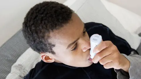 A boy is sat on a sofa or bed breathing on an inhaler. The photo is cropped at his head and shoulders.