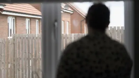 Anonymous image of a mother we are calling Caroline. She has dark hair. Her image is blurred to protect her identity. She is looking though a window into a back garden with a wooden fence and a row of newish houses in the background.