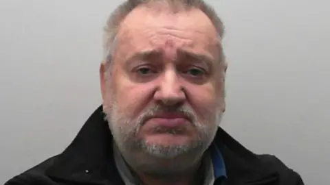 Ian Mark Wrightson has short grey hair and a short grey stubble. He is wearing a black jacket, against a grey background.