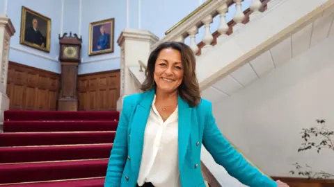 Linden Kemkaran stands on a staircase and holds the bannister with one hand. She is wearing a blue suit jacket and smiling at the camera. Behind her the staircase is covered in a red carpet and a grandfather clock stands on a landing area.