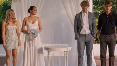 Erika Doss/Prime Two women, one wearing a white dress and holding flowers, stand next to two men with a white table in between