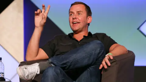 Scott Mills gestures as he speaks onstage wihle wearing a polo shirt, jeans and trainers, sitting down against a colourful backdrop, at the Edinburgh Fringe Festival in August 2014.