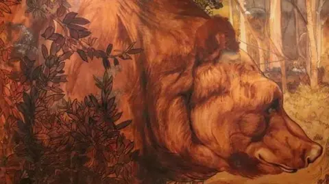 A water colour painting shows a bear in a forest with leaves in front of it. The bear's fur is brown and the forest is sunlit with the tree trunks visible. The bear fills most of the painting and looks sleepy and gentle.