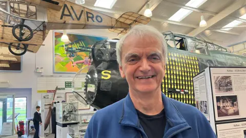 Photograph of Ian Lomax from the Avro Heritage Museum in Woodford. he is pictured in front of the cockpit of a Lancaster Bomber.