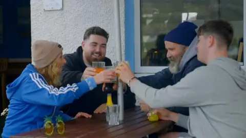 Solus Productions/BBC Jules, clad in a blue jacket and beige hat, clinks her glass in a toast with her son Benny beside her, son Chevy across from her, and husband Greg. They are gathered outside on a picnic table adjacent to a building.