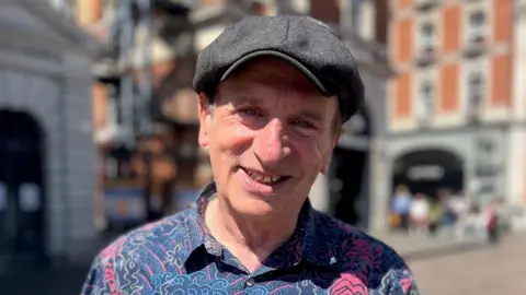 A middle-aged man wearing a baker-boy cap and a patterned shirt in blue and pink, featuring tigers, leaves and swirls. It is sunny and he is smiling.