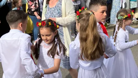 Five children dressed in white with floral head dresses dance through the streets of Helston, watched by a crowd of happy people 