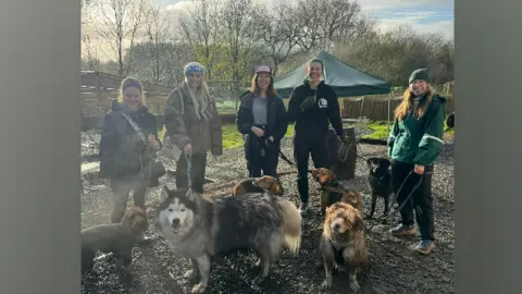 Sarah Reilly Five women smile at the camera, holding dogs on a lead, on a grave path with fields behind them