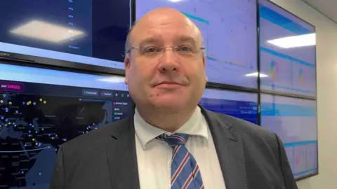 A man standing in front of multiple TVs with different graphs and numbers on them, he is wearing a suit and tie and a pair of glasses.