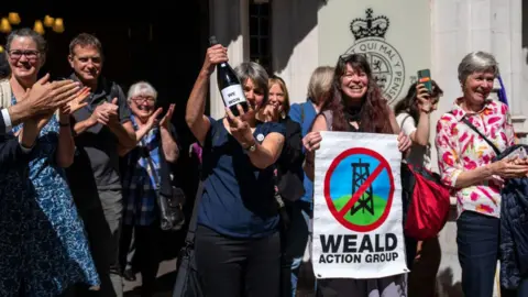 Getty Images A group of conservation activists celebrating outside a Supreme Court. One of them is holding a small banner with the words Weald Action Group. There is also a picture of an oil refinery, drawn inside a red circle, and with a large red cross over it.