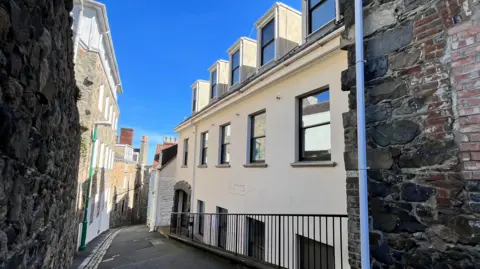 A three-storey office building on a lane in St Peter Port. The image is taken at the top of a hill, looking down. The building is to the right of the image. It is painted cream. The words 'islands insurance' are faintly visible on the wall. The sky is blue. 
