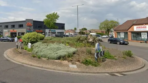 Google A roundabout with green bushes in the centre at the junction of two routes. There are several cars on the roundabout and at the junctions. Two shops are also visible on the junction with houses in the background.