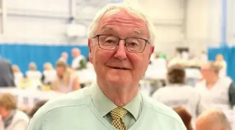 A man with grey hair and a green shirt and pullover with a yellow tie in a room with the blurred shapes of people behind him
