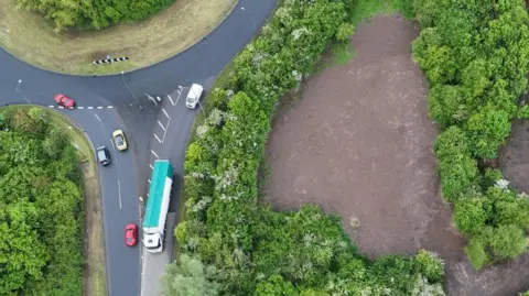 An aerial shot of a large patch of land off a busy roundabout. The patch of land is surrounded by trees. The land has been torn up and there is just brown soil visible now. 