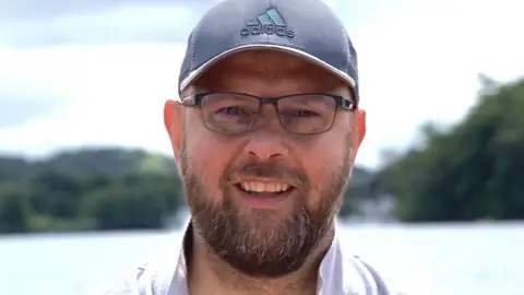 Andrew Slorance has a beard and is wearing glasses and a baseball cap. He is smiling at the camera