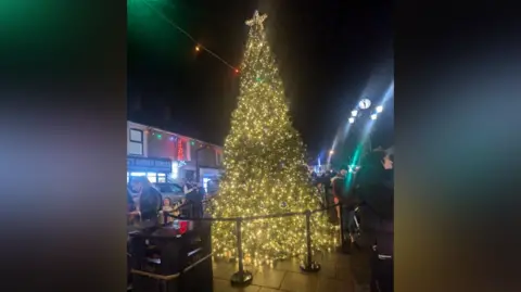 Jade Aubin A night-time scene of a very sparkly Christmas tree with a star on top, lit up. A lightweight webbed cordon is around the tree, with shops lit up in the background