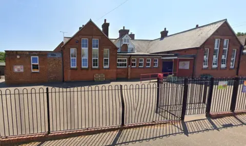 Google External view of Reepham Primary School with its playground visible behind a black metal fence. It is a sunny day.