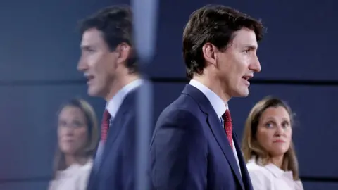 Reuters Justin Trudeau w niebieskim garniturze stoi na pierwszym planie, a Chrystia Freeland w białej koszuli patrzy w jego stronę. Obydwa odbijają się w lustrzanej powierzchni znajdującej się za nimi.  