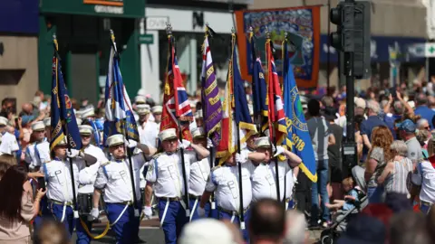 PA Media A number of men are walking and holding flags. It is a sunny day and they are wearing white shirts and hats which have a white top and a black peak. There are crowds in the background.