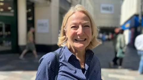 Caroline Wright smiles, she has mid-length blonde hair. She wears a blue and white spotted shirt. You can see a bag strap over her shoulder.