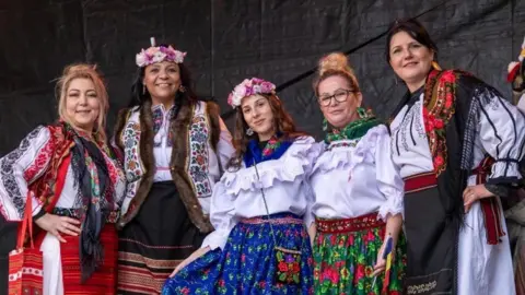 Five women are stood in a row dressed in traditional Romanian clothes. They are wearing brightly coloured flowered skirts, white frilly blouses, fringed shawls and fur lined gillets and some have pink and white floral headdresses