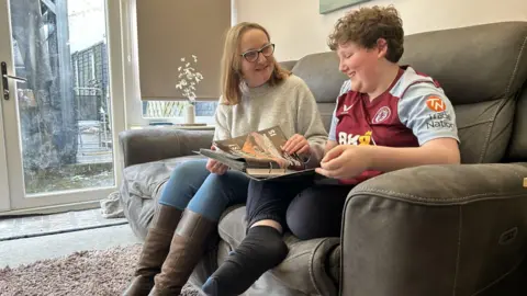 Amy wears glasses, a gray jumper and long brown shoes. She is sitting next to her 12 -year -old son Cooper, a child who has brown hair and is wearing Aston Villa Top. They are sitting on a couch in a living room, looking at a French door book behind them. They are both smiling.