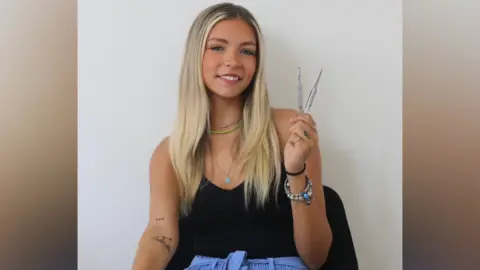 Kiera Robinson is smiling at the camera, has blonde hair and blue eyes. She is wearing a black strappy top, blue trousers and is holding two pairs of tweezers.