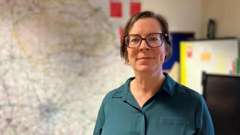 BBC/Seb Cheer A woman, Rachel Joyce, looks into the camera. She is standing in an office-style room with a map behind her, showing the North York Moors.