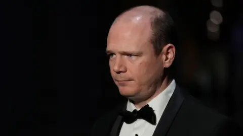 A man in a tuxedo. He has hair on the sides of his head. The background is black.
