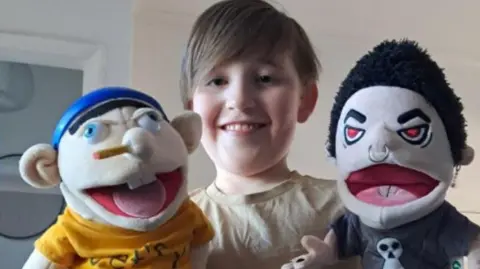Leighton smiling at the camera with a beige t-shirt, holding his two puppets in either hand.