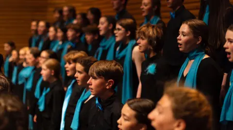 Members of the Farnham Youth Choir stand and sing in unison while performing to a crowd. They are positioned in rows, elevated from front to back. They are all wearing black shirts with a variation of teal blue scarves, neckties or pins.