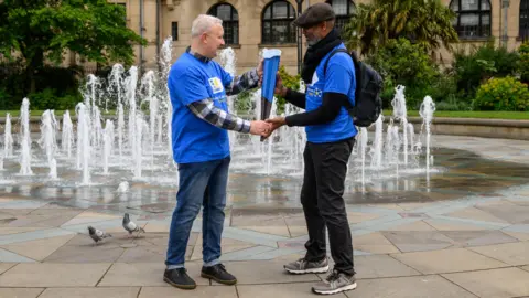 Yorkshire Cancer Research Two men passing the baton to each other in a city square, which has fountains and a few pigeons in the background. They wear blue t-shirts. 