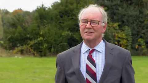 BBC A man wearing glasses and a gray suit with a blue and white criss-cross shirt and a red, navy and white striped tie. He is standing outdoors with an area of ​​grass and trees in the background.