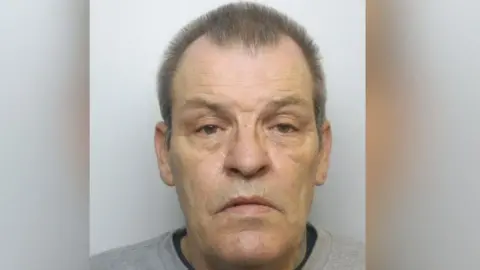 Sydney Burdon looking serious at the camera for his mug shot picture. He has a receding hairline and brown hair, wearing a grey top on. 