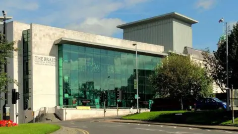 The exterior of the Braid building in Ballymena. The modern grey stone building has a large glass frontage and glass panels at the side.  There is a lawned area to the front with a few trees and two sets of traffic lights are on the road leading to the entrances