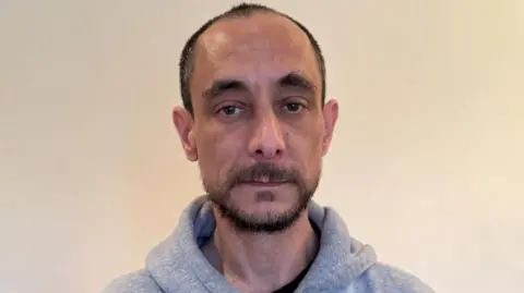 Steve looks at the camera and wears a grey hooded jumper. He has a dark beard, brown eyes and is partly bald. The background is blank.