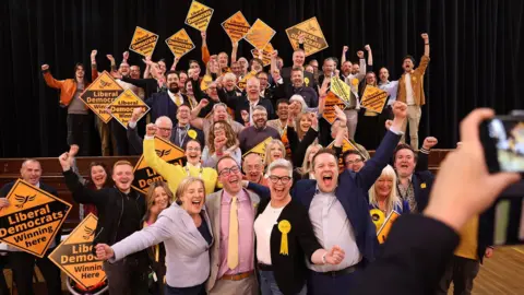 Sean Hansford A group of Lib Dem councillors leap in the air in delight - holding orange signs saying Liberal Democrats winning here