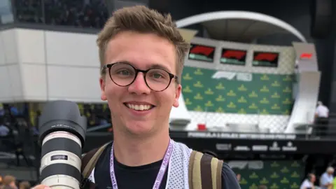 Craig Evans at a racing event holding his camera. He has spiky light brown hair and round glasses. He is stood in front of a crowd of people and he has a lanyard around his neck