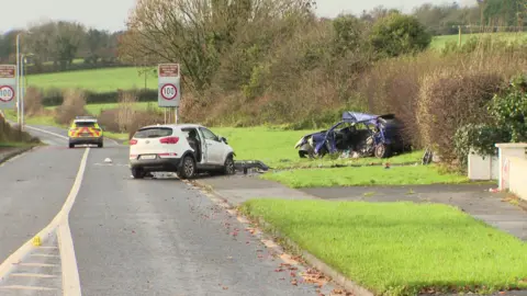 Two cars are vislble. A blue car is on the grass. One side of the vehicle is removed and the inside is visible including the steering wheel and rear seats. The roof is partially off the car and debris is strewn around the vehicle. A second white vehicle is on the road.