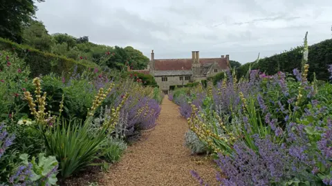 fiona A gravel path runs between two large flower beds full of purple and yellow flowers. At the end of the path is a grey stone building that has red tiles on its roof.