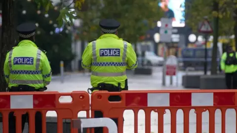 Two police officers in florescent shirts lean against a road barrier.  