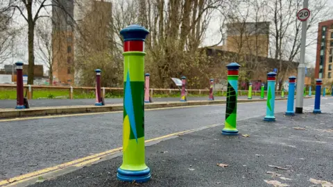 Upfest Image shows bollards flanking a road. They are painted in geometric shapes in varying greens and blues. There is a small patch of trees in the background and some blocks of flats