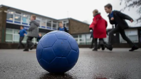 A blue football sits on a school playground. Primary school pupils can be seen running around in the background wearing winter coats. A two-storey school building is behind them.  