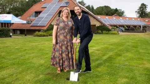 Two people stand on a neatly cut lawn in front of a modern building with a red‑tiled roof covered in solar panels. One of them holds a shovel, taking part in an event.