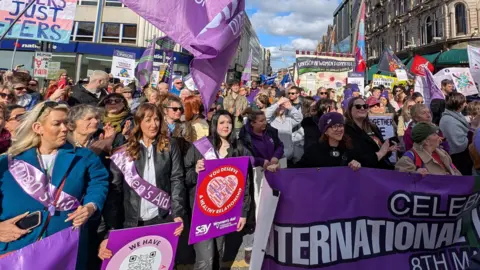 Women standing together holding purple posters, banners and flags as they take part in a rally.