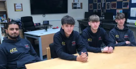 The Brickie Boys sit around a table, wearing dark hoodies with a ACE logo in yellow top left.