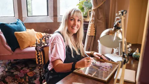 BBC/DSP/Georgina Vincent A woman sat at a desk painting on a mirror while smiling into the camera