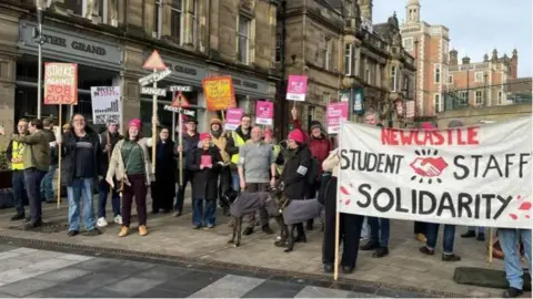 Men and women line a town square pavement, holding up posters, banners and placards that read 'strike against job cuts' and 'strike picket line'. There is a row of buildings behind them. 